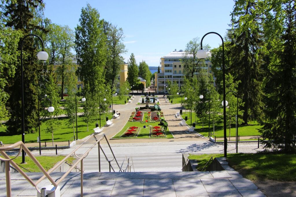 Portaiden yläpäästä otettu kuva puistosta. Keskellä kukkaistutuksia ja penkkien reunustamat hiekkatiet. Vasemmalla ja oikealla puista puistoaluetta. Edessäpäin rakennuksia ja niiden takana pilkistää järvi.