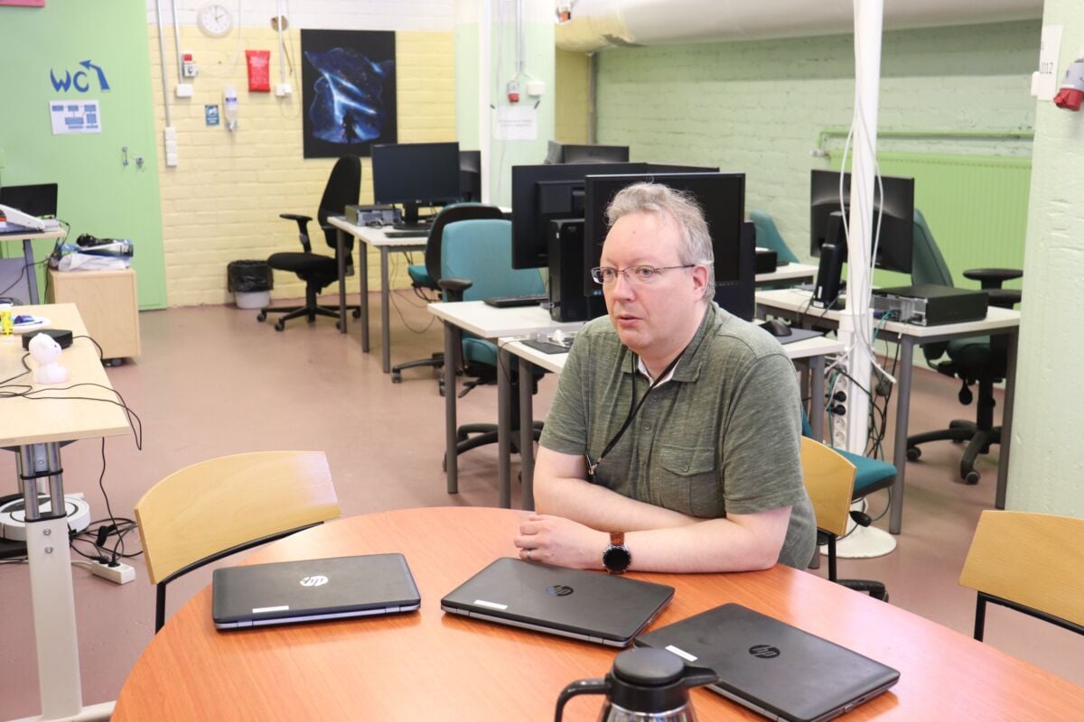 Tietokoneluokka jossa työskentelypöytyiä' ja tietokoneita. Kuvan etutalalla mies istuu pöydän ääressä, pöydällä kolme kannettavaa tietokonetta.