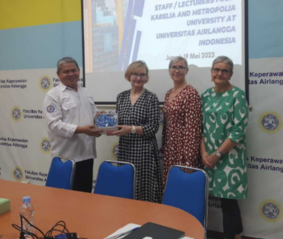 Neljä henkilöä seisoo rinnakkain valkokankaan edessä. Seinällä teksti Karelia and Metropolia university at University Airlangga Indonesia